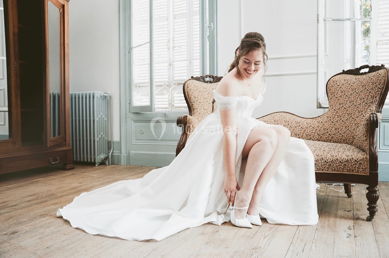 Une femme en robe de mariée ajuste ses chaussures assise sur un canapé dans une pièce lumineuse.