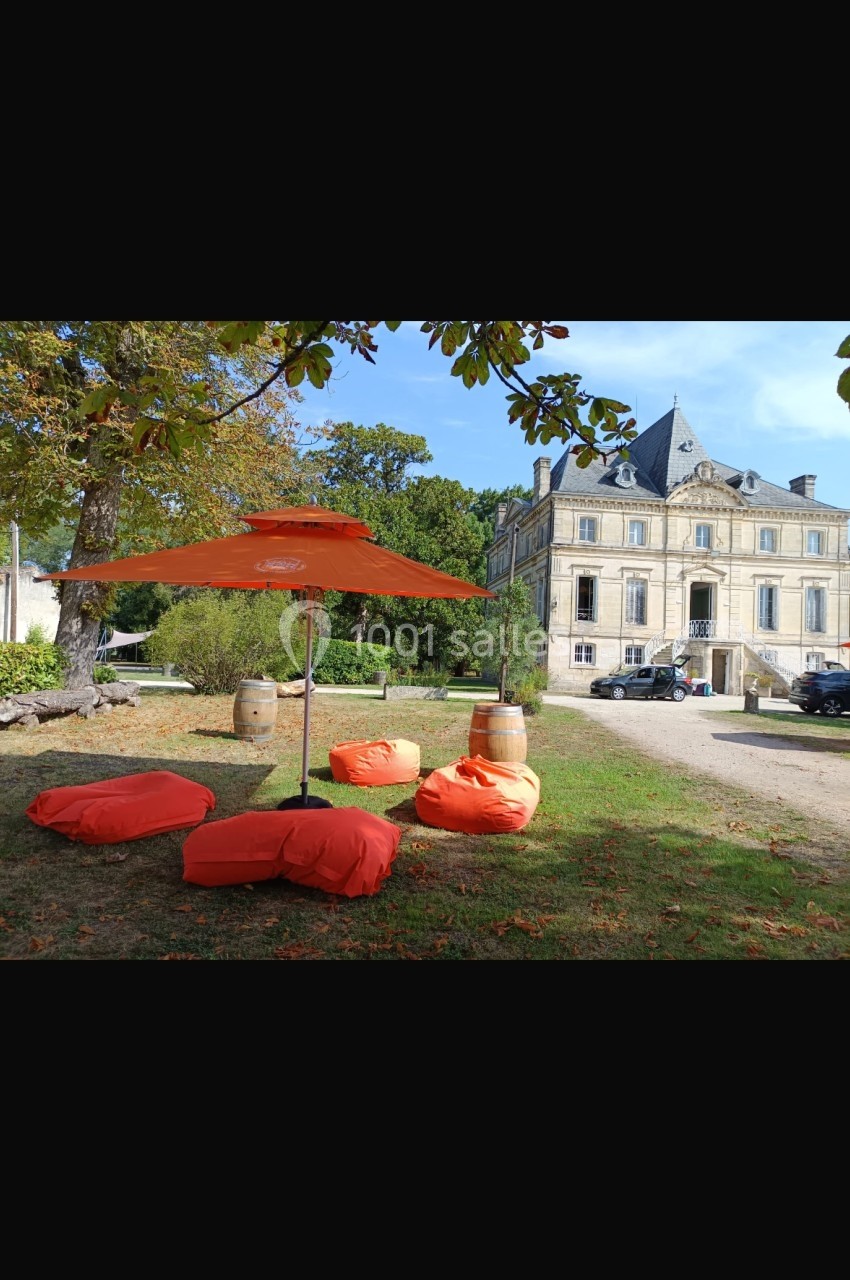 Chaises et coussins orange avec parasol sur une pelouse devant un bâtiment ancien entouré de verdure.