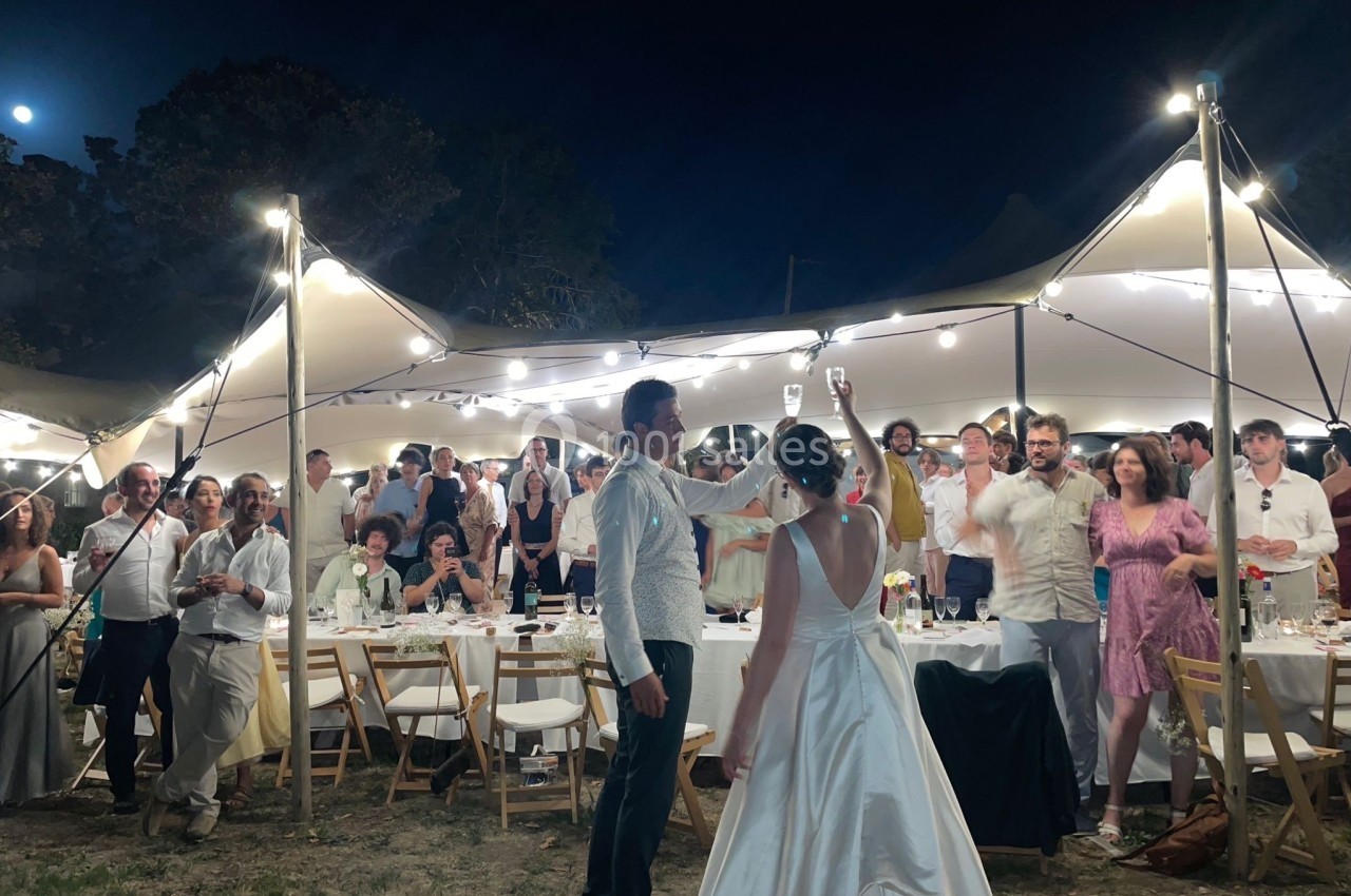 Un couple en tenue de mariage lève un verre devant des invités assis et debout sous des guirlandes lumineuses en extérieur.
