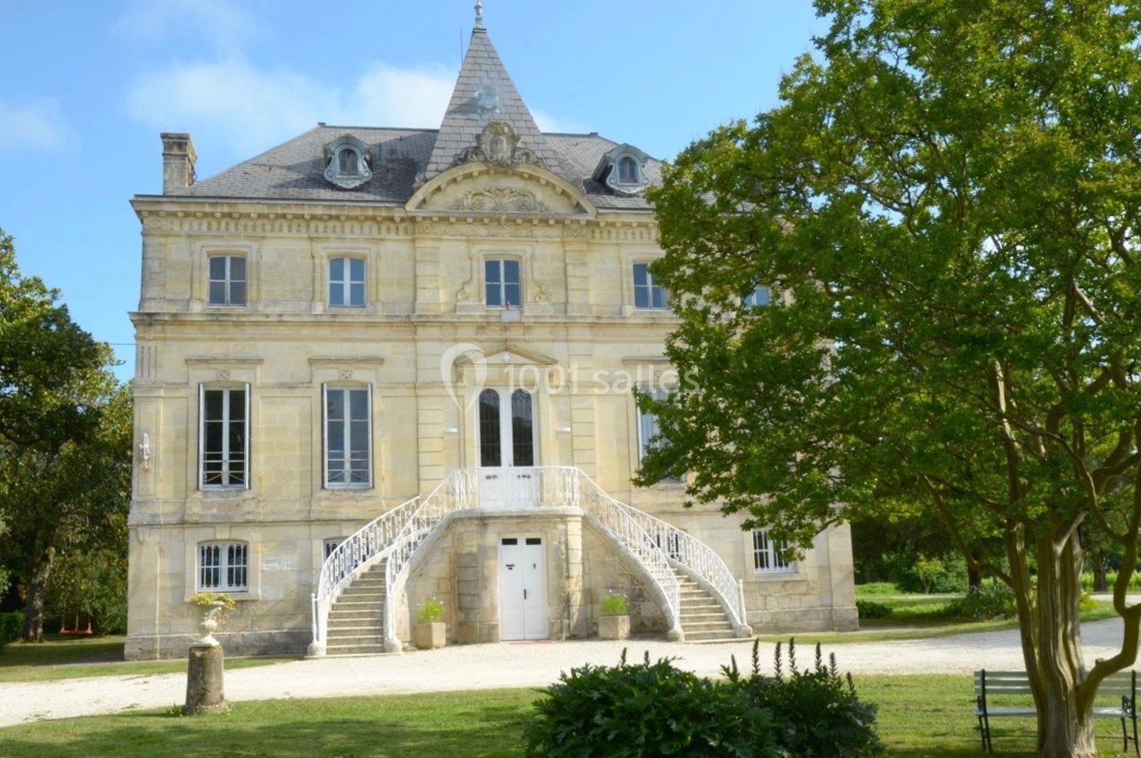 Façade d'un manoir en pierre avec escalier central, entouré de verdure et d'arbres sous un ciel dégagé.