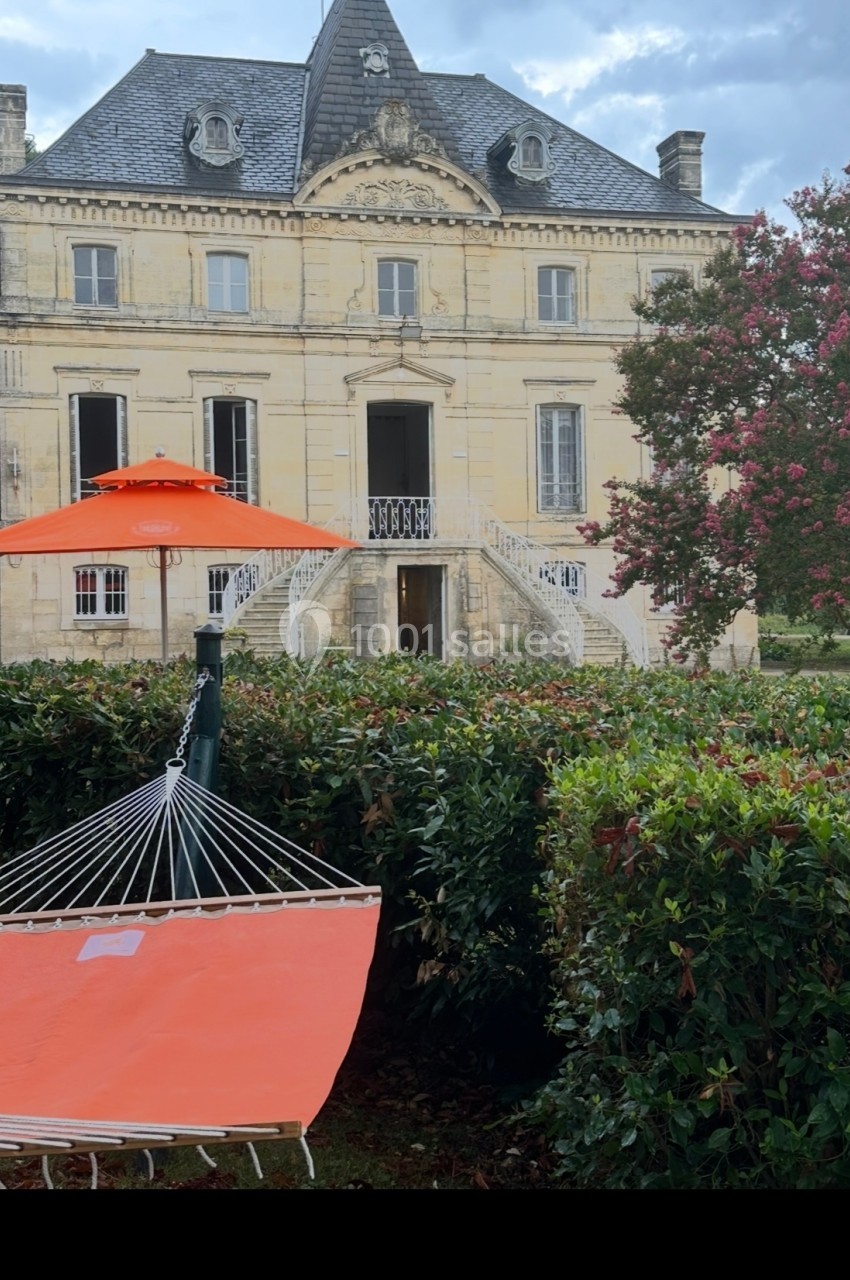 Hamac orange installé dans un jardin devant une grande maison en pierre avec un toit mansardé et des escaliers.