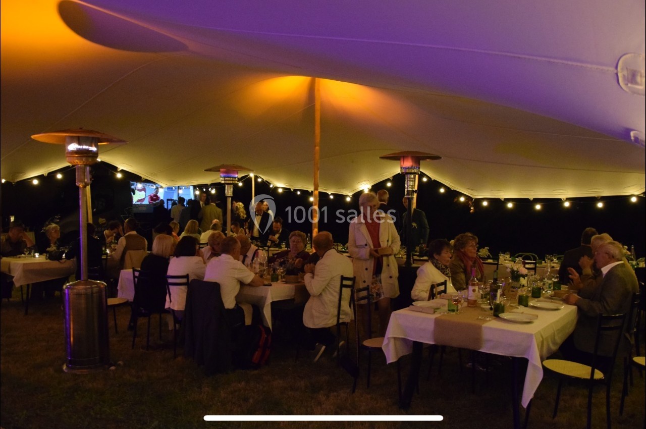 Des convives dînent sous une grande tente éclairée, avec des tables dressées et des guirlandes lumineuses.