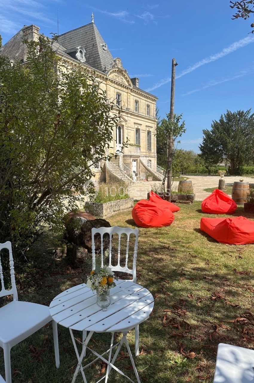 Table et chaises blanches sur une pelouse devant un bâtiment ancien, avec des poufs rouges et des arbres en arrière-plan.