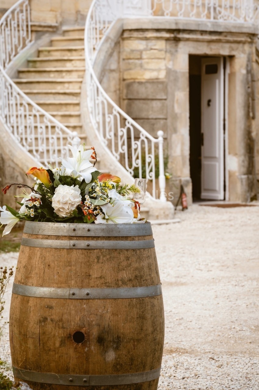 Fût en bois décoré de fleurs blanches et colorées, placé devant un escalier en pierre et une porte ouverte.