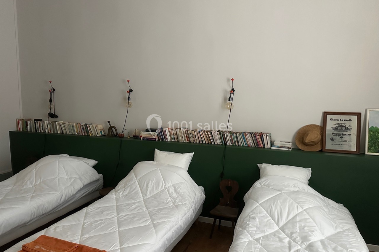 Trois lits simples alignés avec des couvertures blanches, une étagère murale avec des livres et des lampes suspendues.