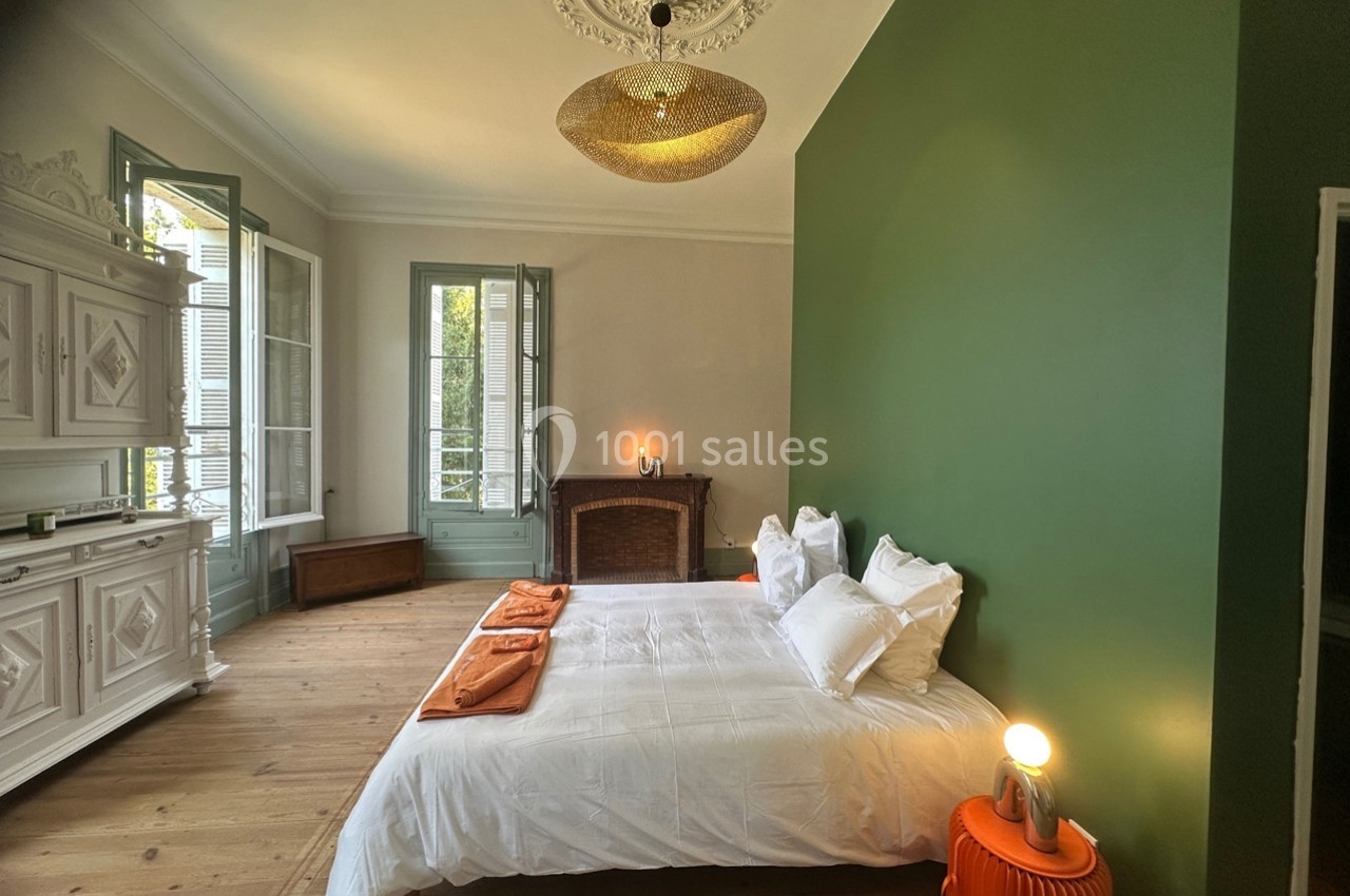 Chambre lumineuse avec murs verts, lit double, cheminée ancienne, mobilier blanc et grandes fenêtres ouvertes.