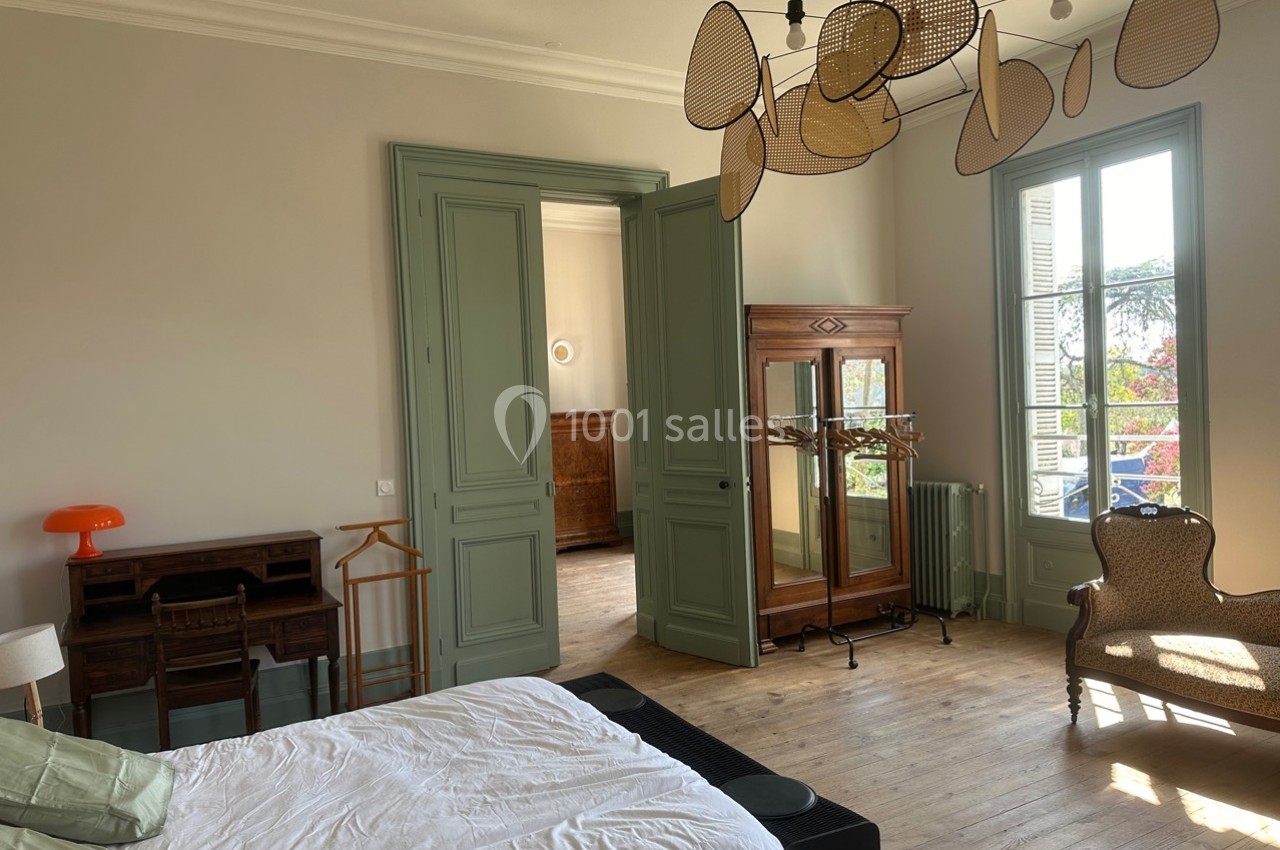 Chambre lumineuse avec lit, bureau ancien, armoire en bois, fauteuil et grande fenêtre donnant sur un extérieur arboré.