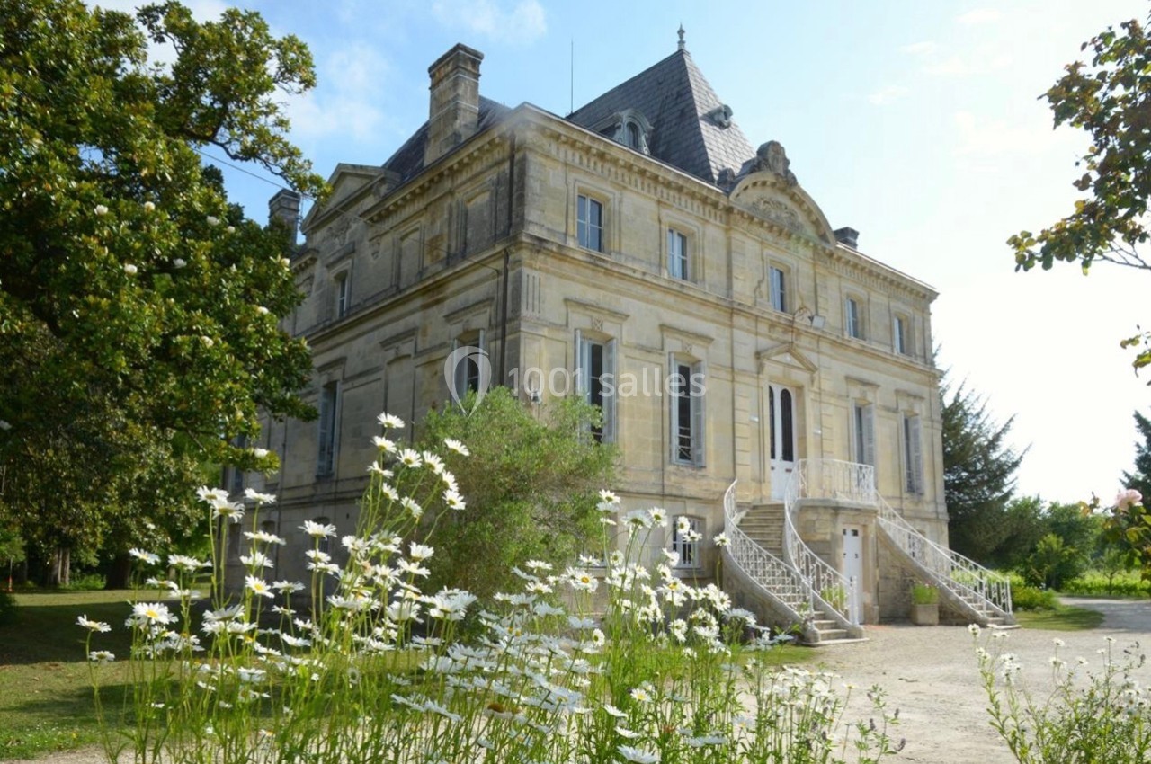 Façade d'un château en pierre entouré de verdure, avec des marguerites au premier plan.