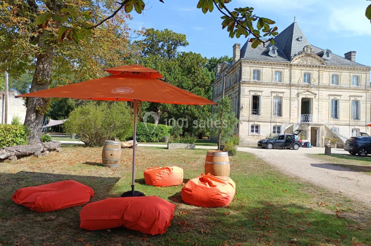 Espace extérieur avec poufs et parasol orange devant un bâtiment ancien entouré de verdure.