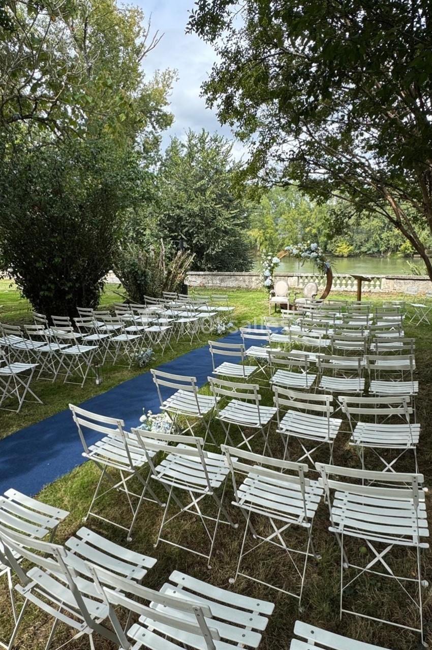 Chaises blanches alignées en extérieur, disposées face à une arche décorée pour une cérémonie près d'un cours d'eau.