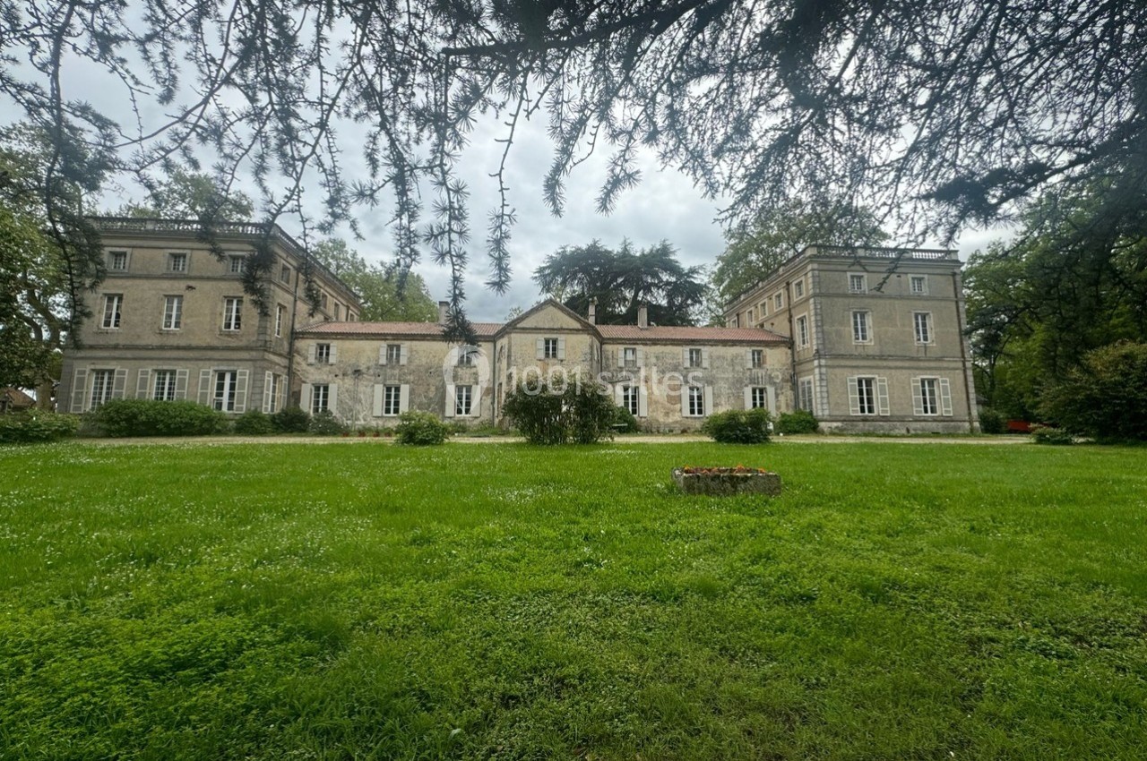 Façade d'un grand bâtiment ancien entouré de pelouses et d'arbres, sous un ciel nuageux.