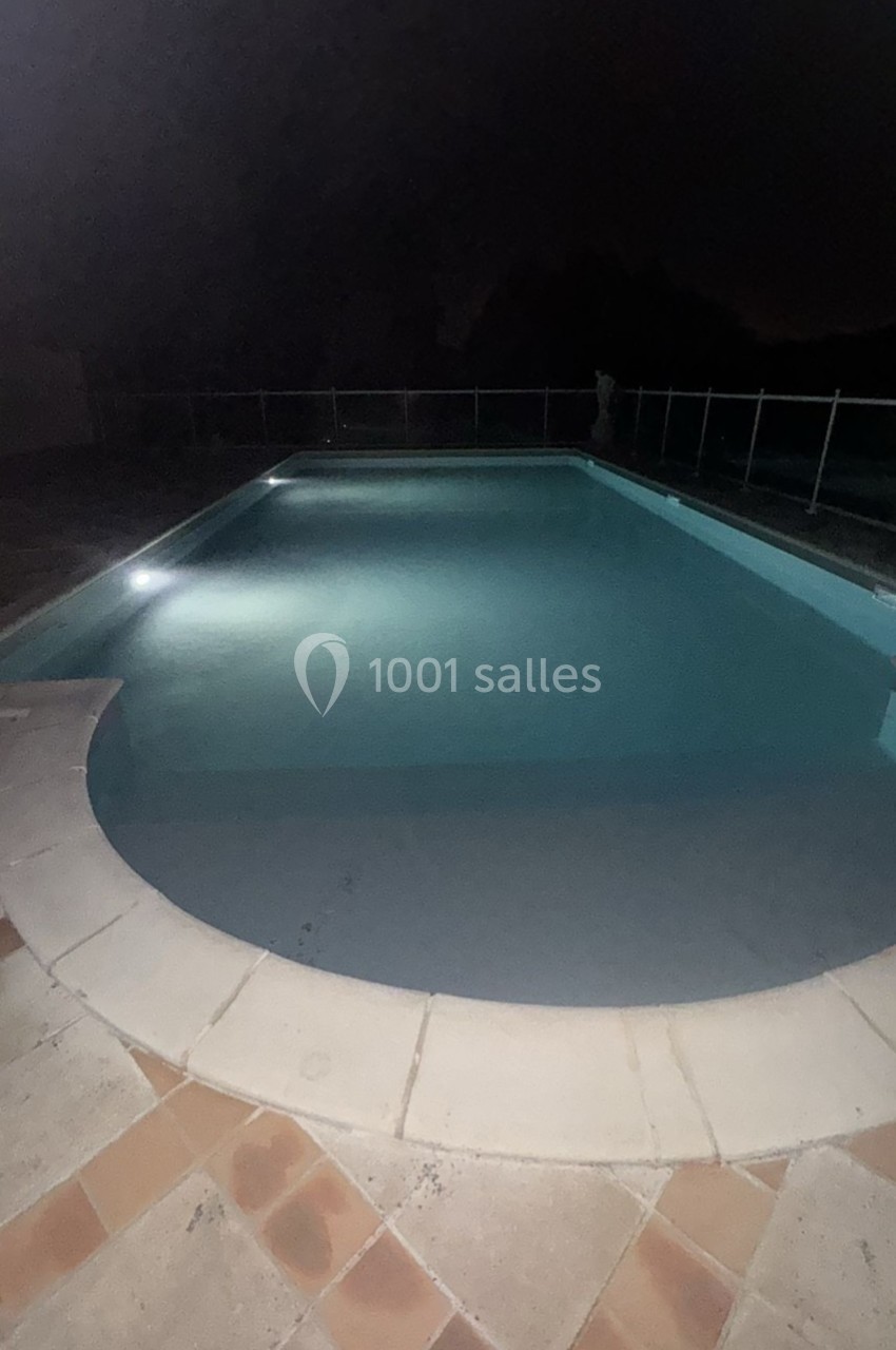 Piscine éclairée la nuit avec margelle en pierre et carrelage au sol, entourée d'une barrière de sécurité.