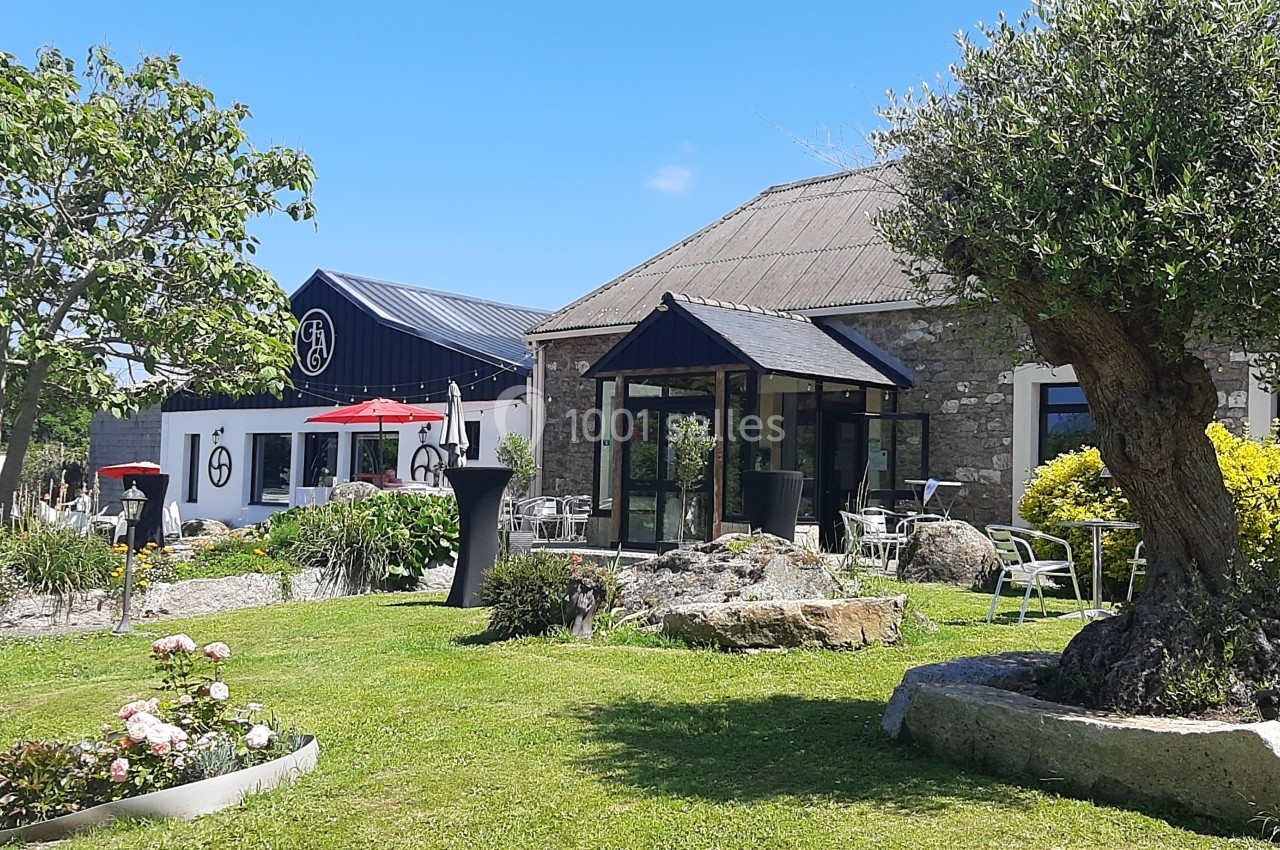 Extérieur d'un bâtiment en pierre avec terrasse, parasols rouges, jardin fleuri et pelouse sous un ciel bleu.