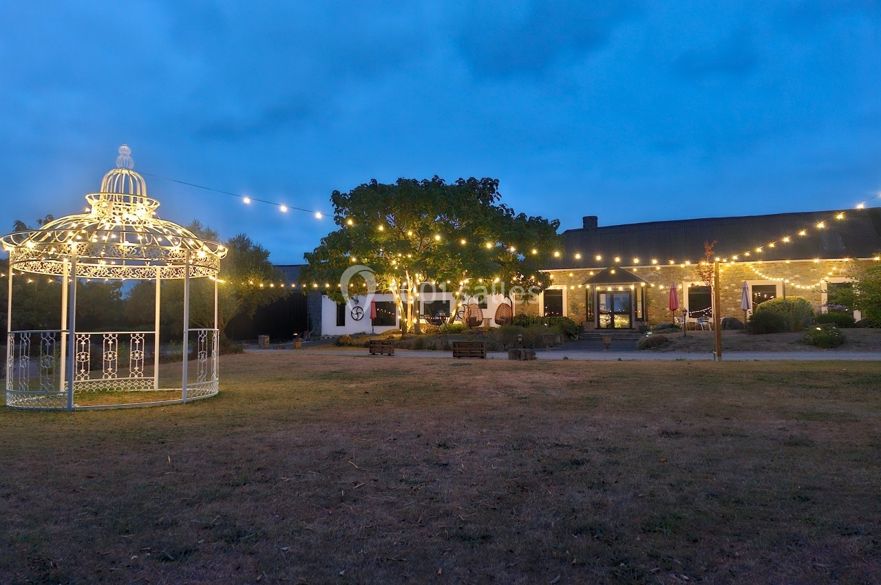 Jardin éclairé au crépuscule avec un kiosque illuminé et des guirlandes lumineuses suspendues près d'un bâtiment.