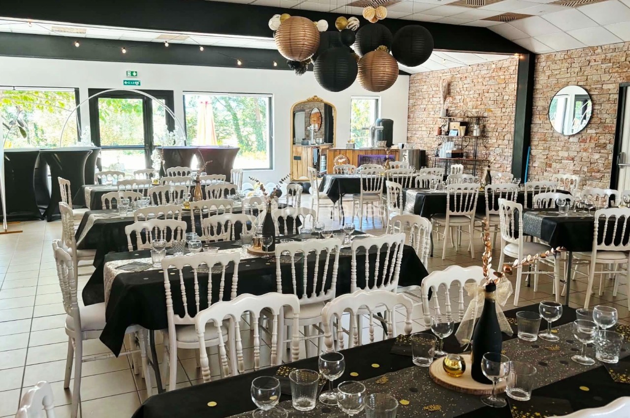 Salle de réception décorée avec des tables dressées en noir et or, chaises blanches et lanternes suspendues.