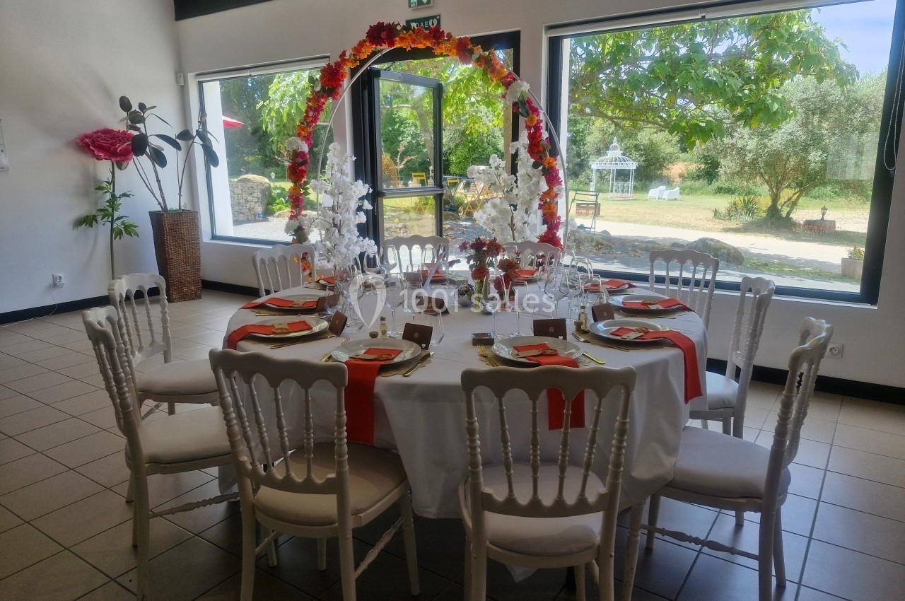Table ronde décorée pour un événement, avec nappes blanches, chaises blanches et arche florale, dans une salle lumineuse.