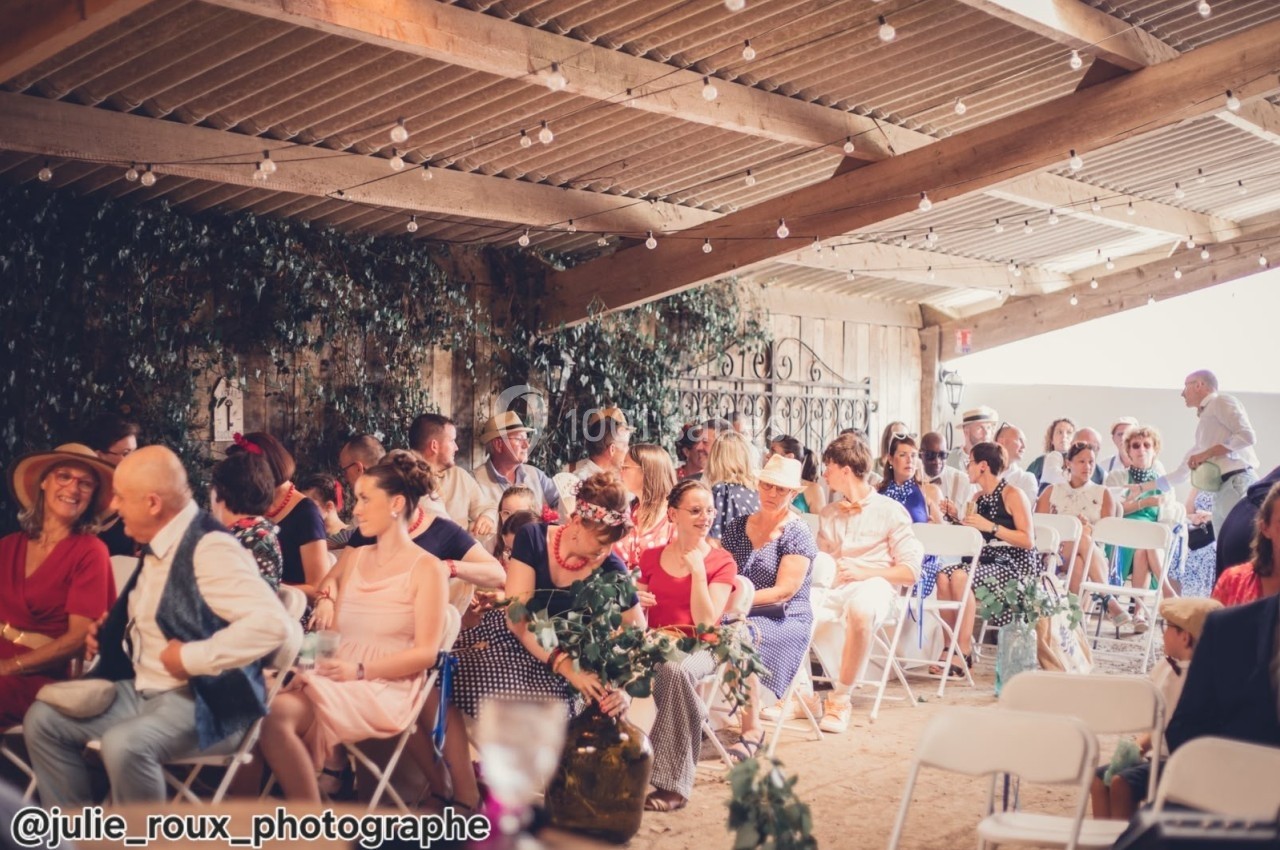 Des invités assis sous une structure en bois décorée de feuillages, lors d'une cérémonie ou d'un événement festif.