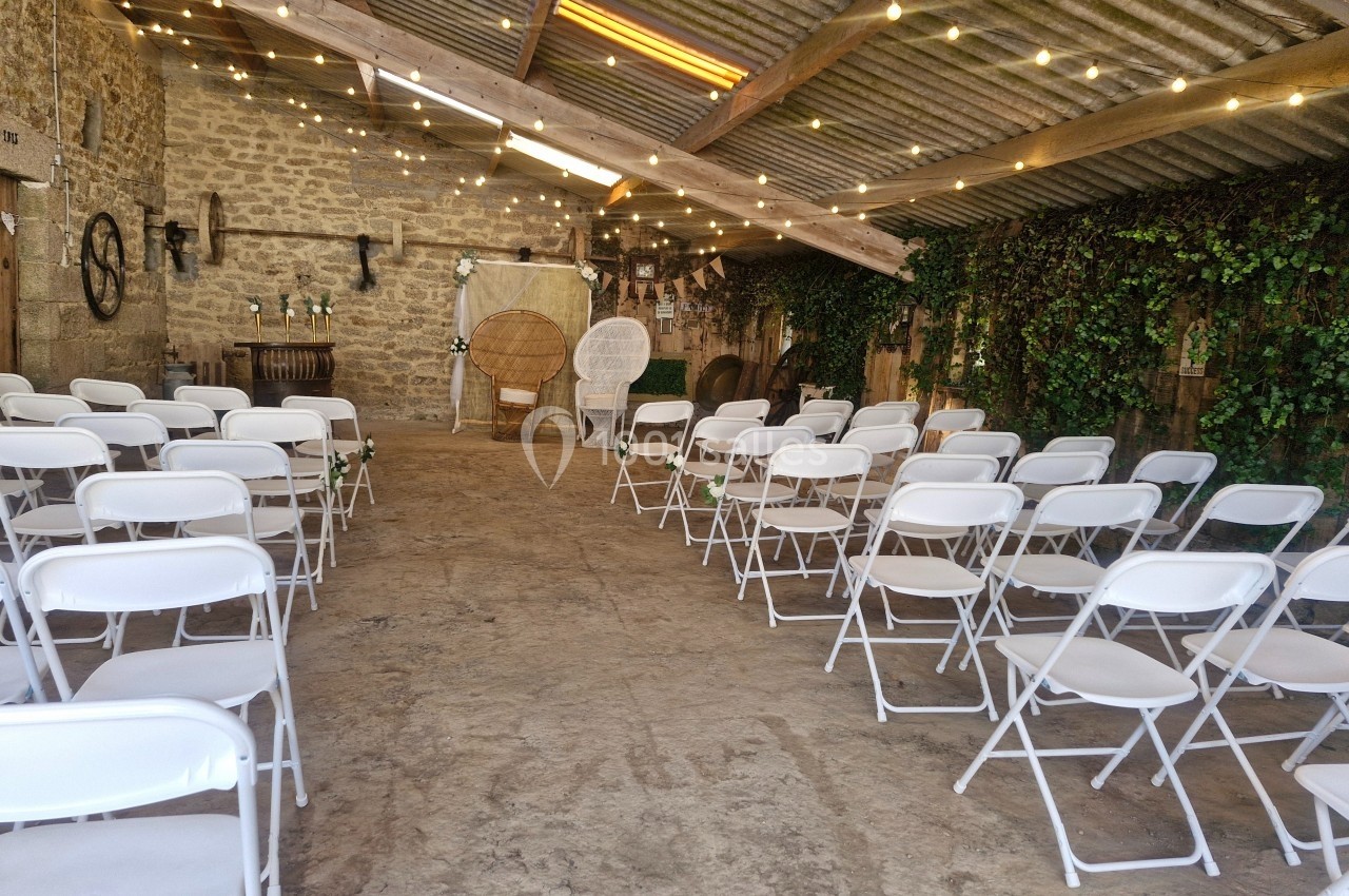 Salle rustique décorée pour un événement avec des chaises blanches alignées, guirlandes lumineuses et murs en pierre.