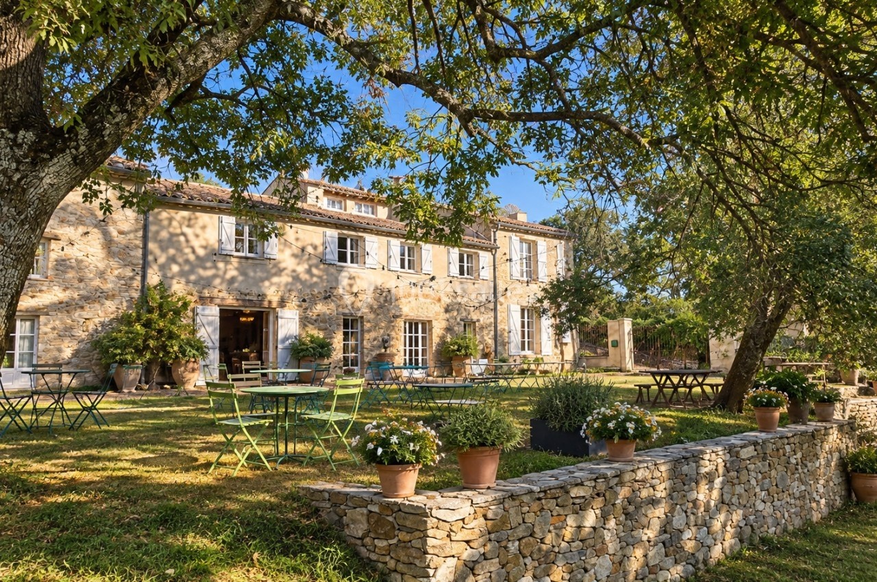 Façade d'une maison en pierre entourée d'arbres, avec des tables et des chaises disposées sur une pelouse ensoleillée.