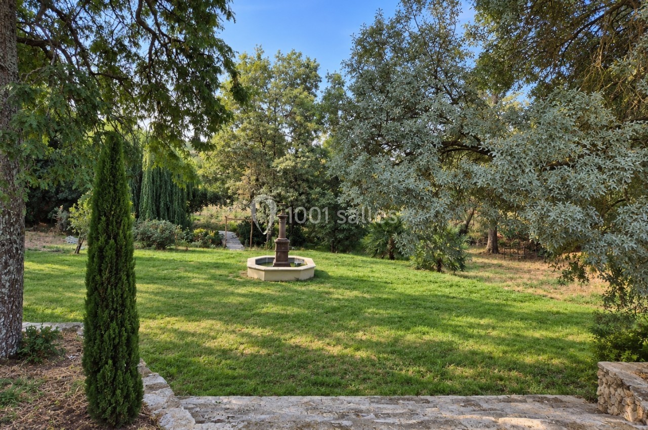Jardin verdoyant avec une fontaine centrale entourée d'arbres et d'arbustes sous un ciel dégagé.