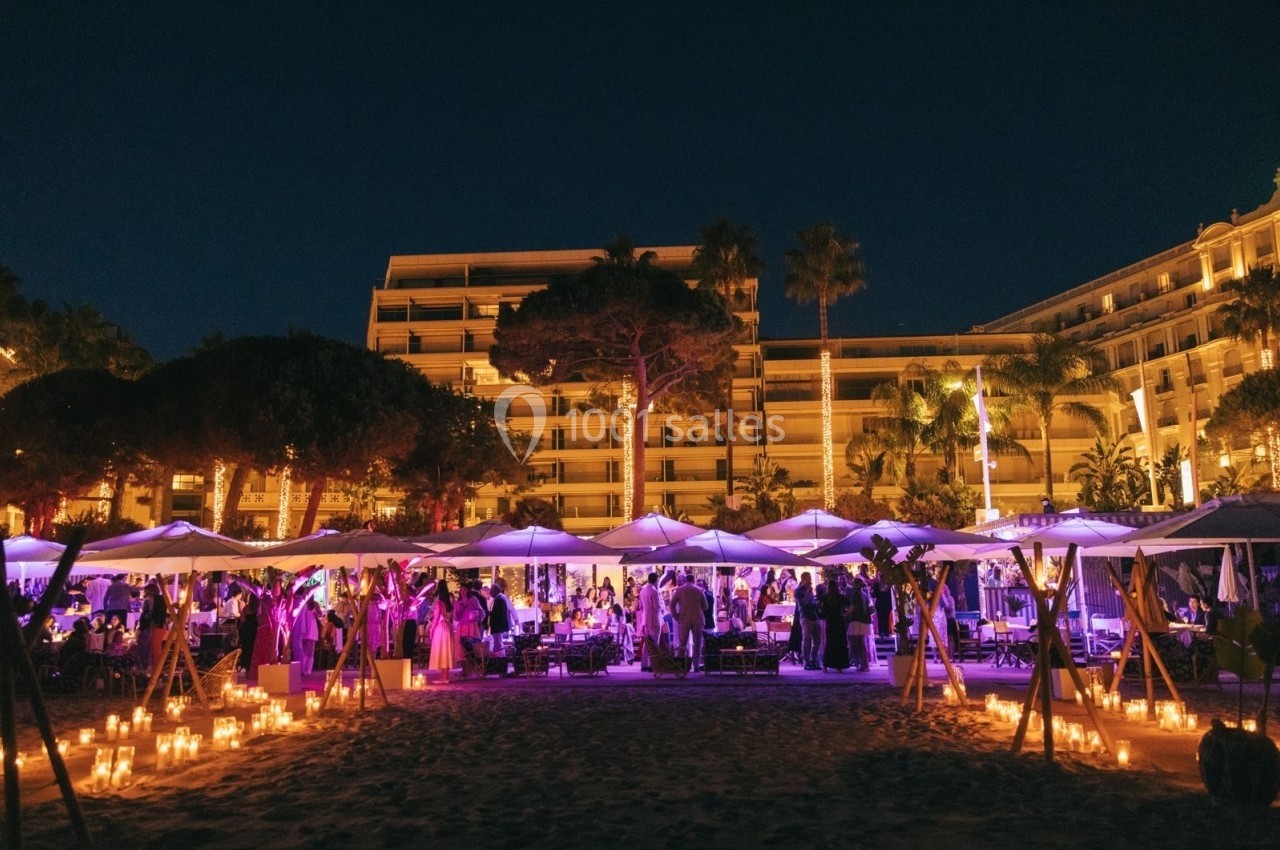 Soirée en plein air sur une plage éclairée par des bougies, avec des invités et des parasols sous un ciel nocturne.