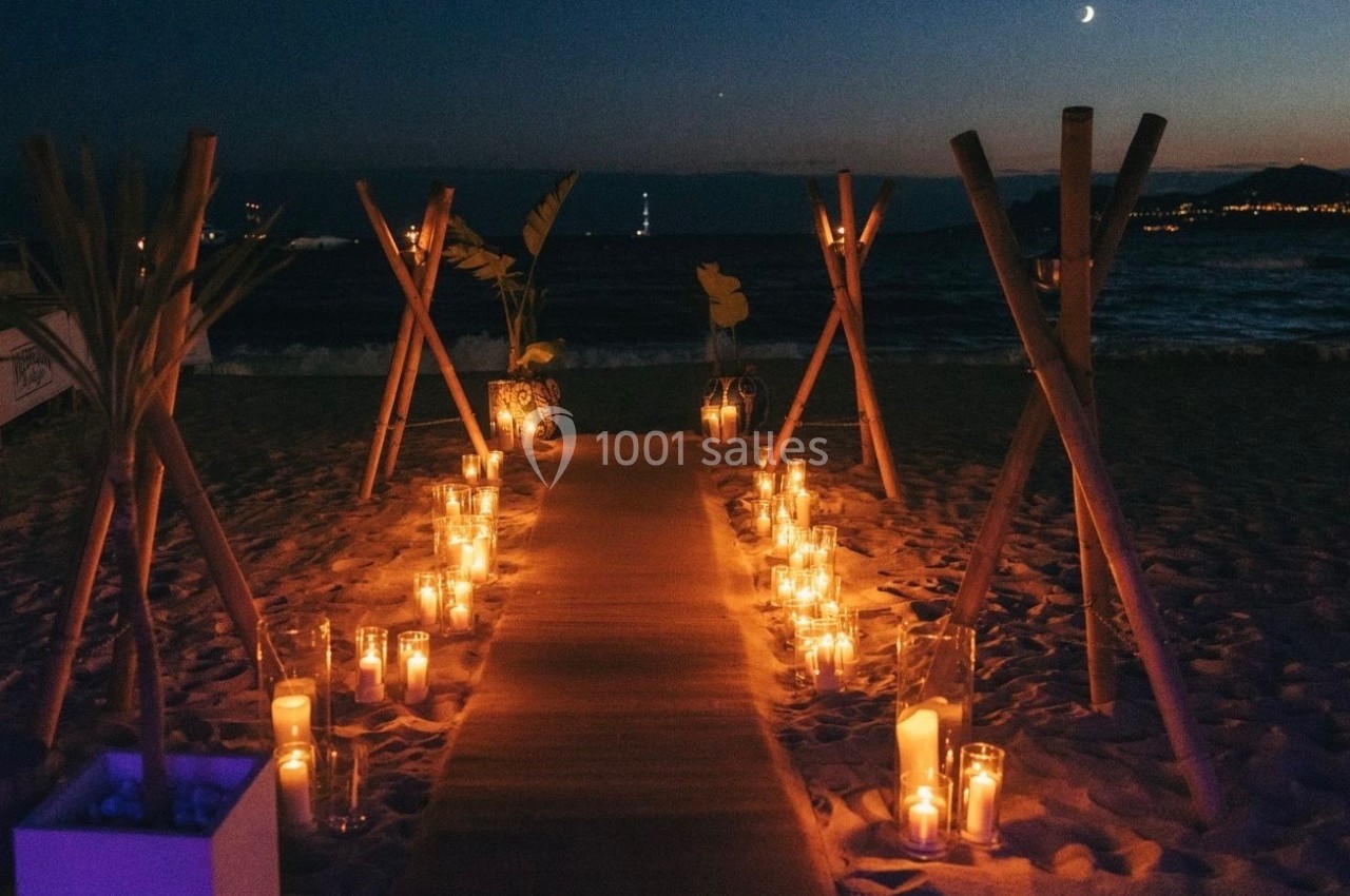 Allée éclairée par des bougies sur une plage au crépuscule, bordée de bambous et de plantes décoratives.