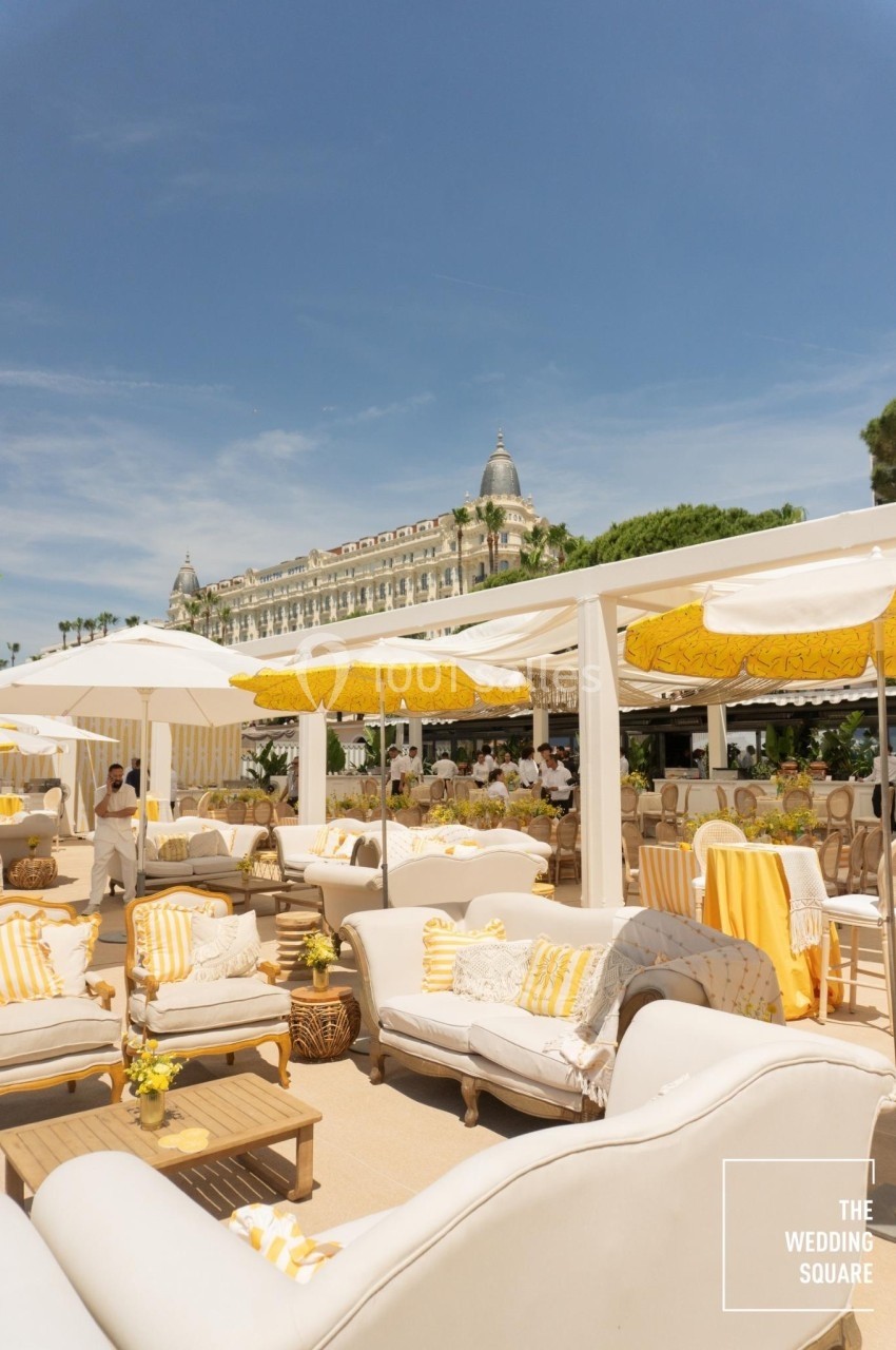 Terrasse ensoleillée avec canapés blancs et jaunes, parasols et vue sur un bâtiment élégant en arrière-plan.