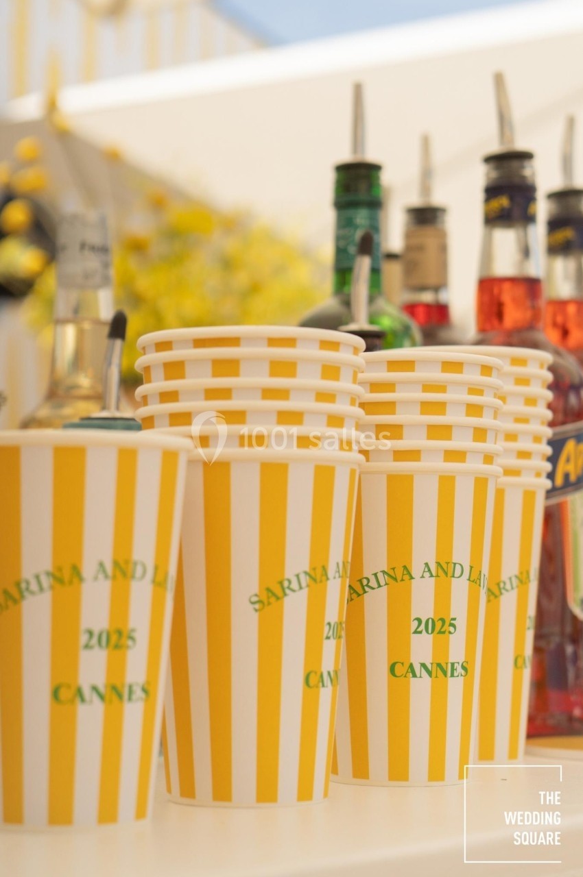 Gobelets rayés jaunes et blancs personnalisés avec des inscriptions, posés devant des bouteilles sur un comptoir.