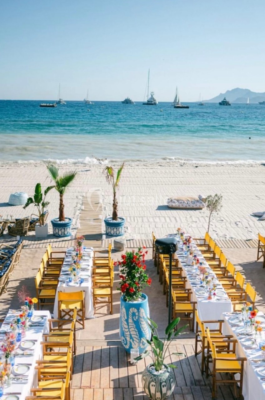 Tables dressées avec des nappes blanches et chaises jaunes sur une terrasse en bois face à une plage et la mer.