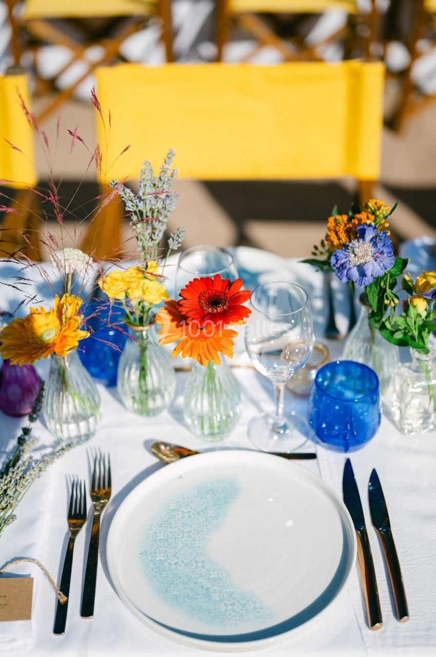 Table dressée avec assiette blanche, couverts, verres bleus et petits bouquets de fleurs colorées.