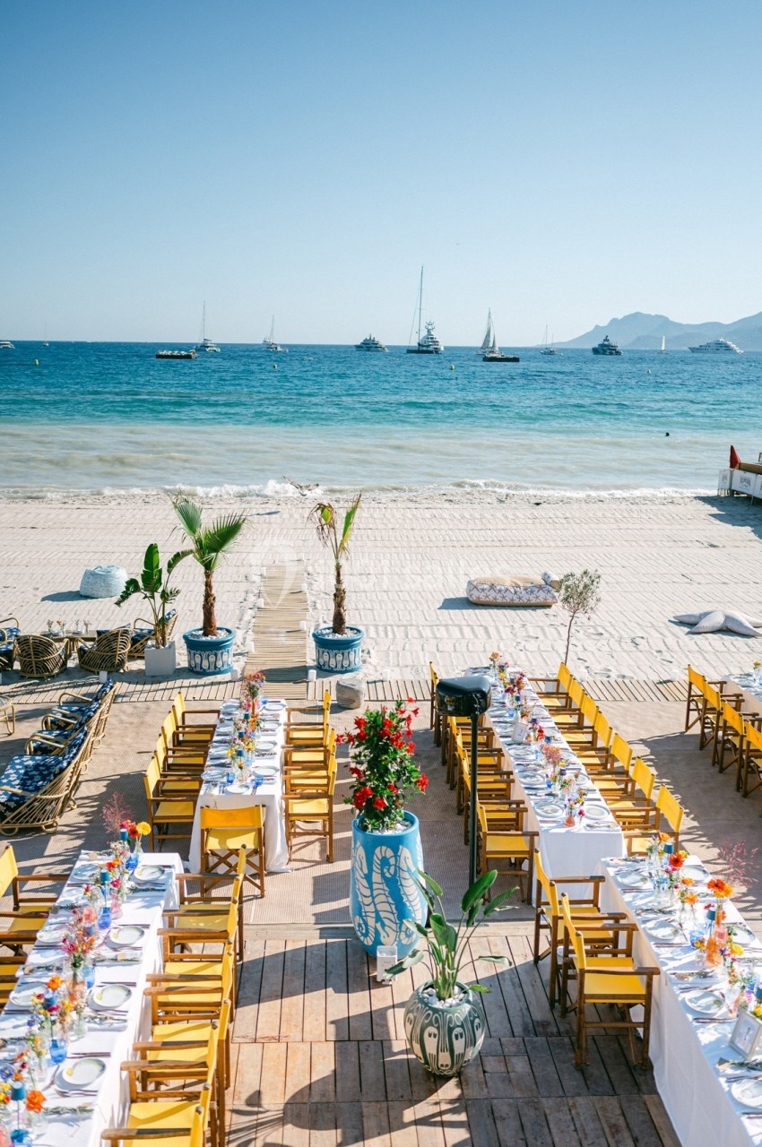 Tables dressées avec des chaises jaunes sur une terrasse en bois face à une plage et une mer calme avec des voiliers au loin.