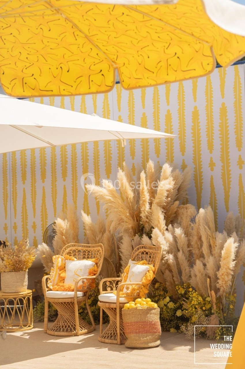 Deux fauteuils en rotin avec coussins jaunes, entourés de plantes séchées, sous des parasols jaunes et blancs.