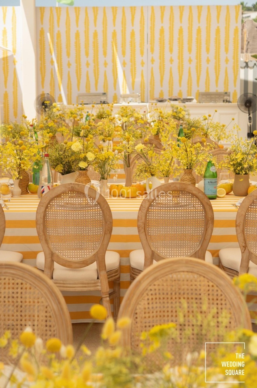 Table décorée avec des fleurs jaunes, nappes rayées jaunes et blanches, et chaises en rotin dans un cadre lumineux.