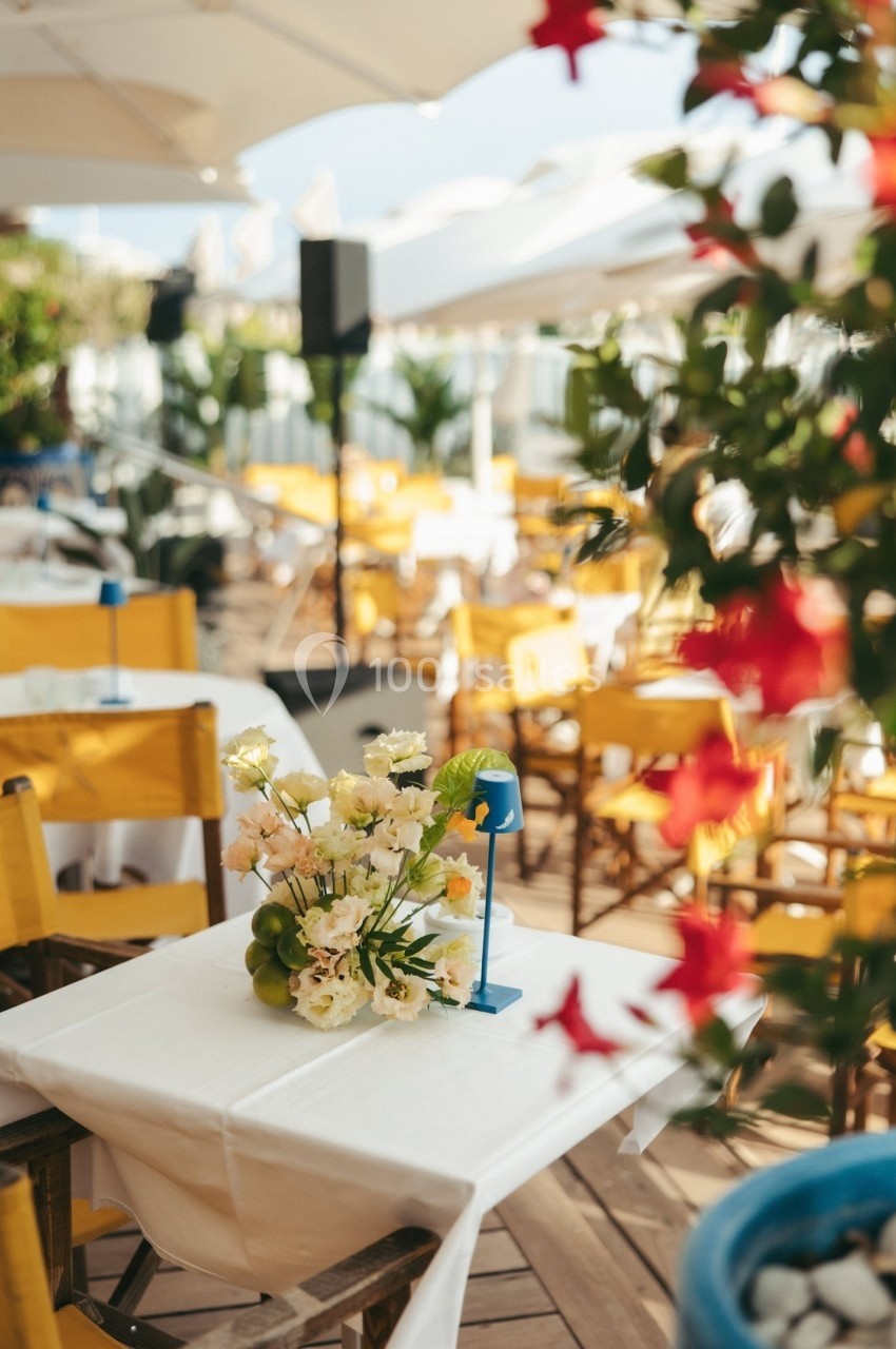 Terrasse ensoleillée avec tables dressées, chaises jaunes, fleurs en centre de table et parasols blancs.