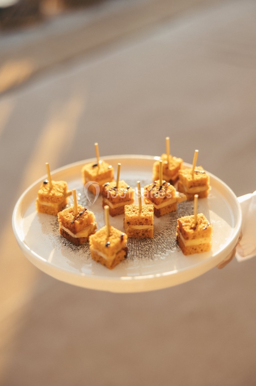 Plateau de bouchées apéritives carrées surmontées de piques, présenté sur un fond flou et lumineux.