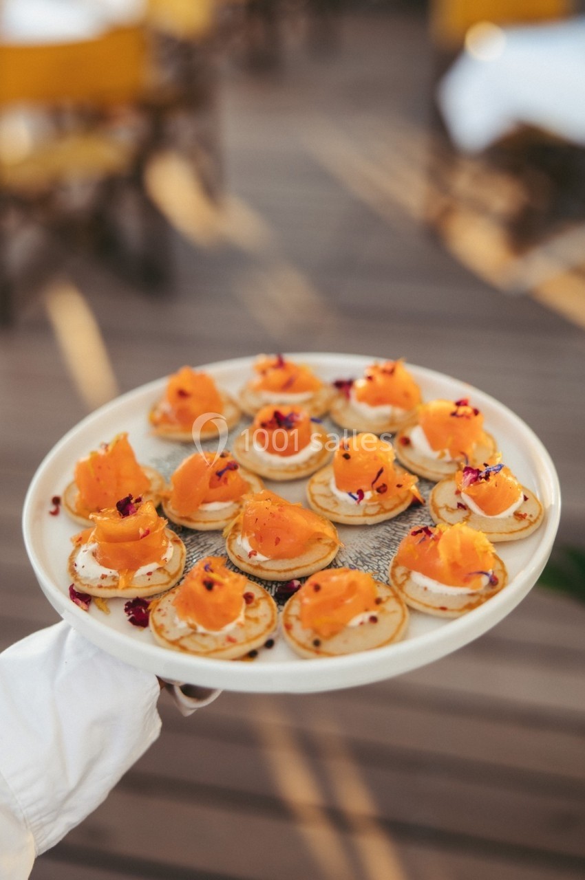 Assiette de blinis garnis de saumon fumé, crème et fleurs comestibles, tenue par une main sur une terrasse ensoleillée.