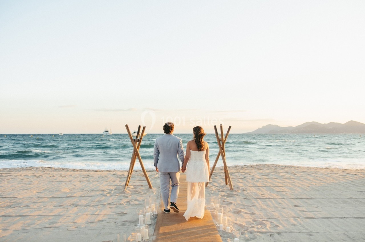 Un couple marche main dans la main sur une plage au coucher du soleil, face à la mer et à une arche en bois.