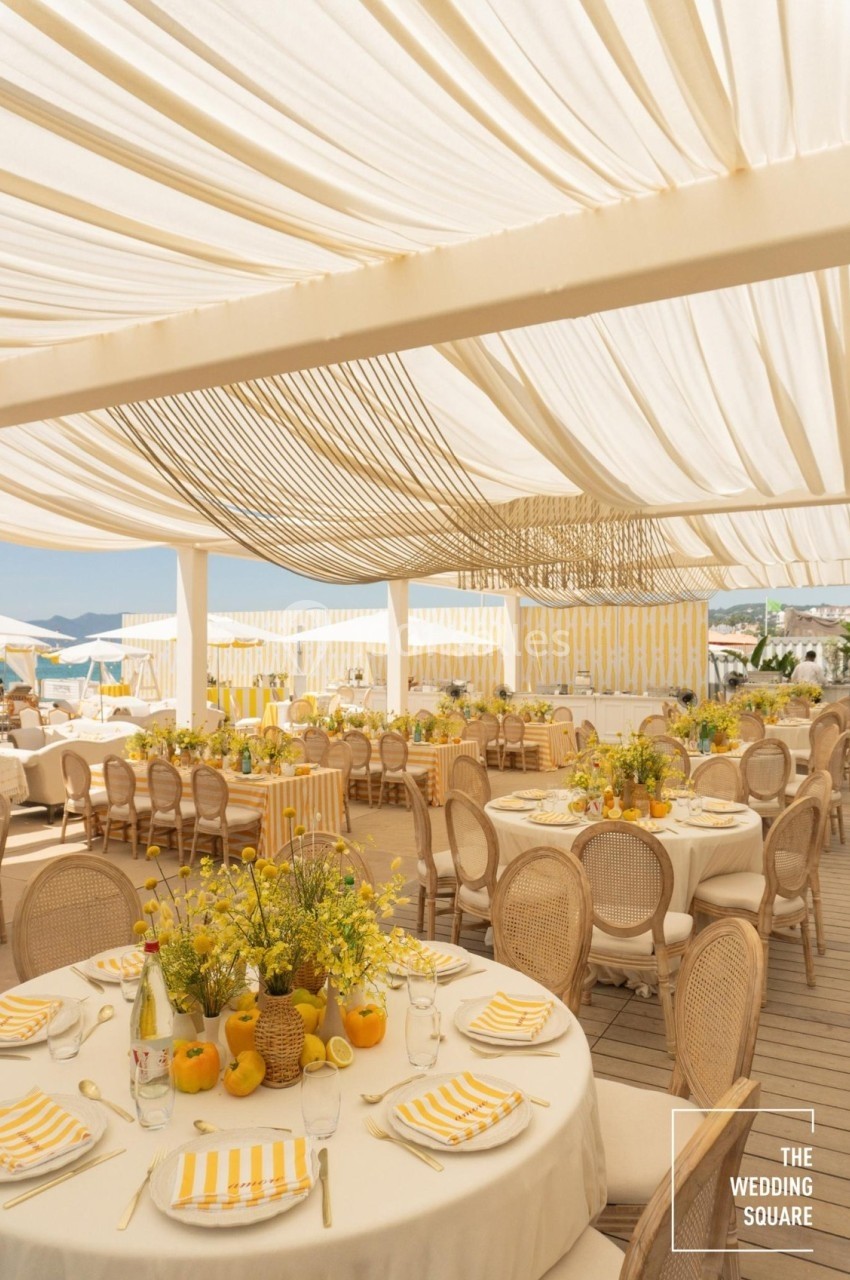 Salle de réception en extérieur avec tables rondes décorées de fleurs jaunes et citrons sous une pergola blanche.
