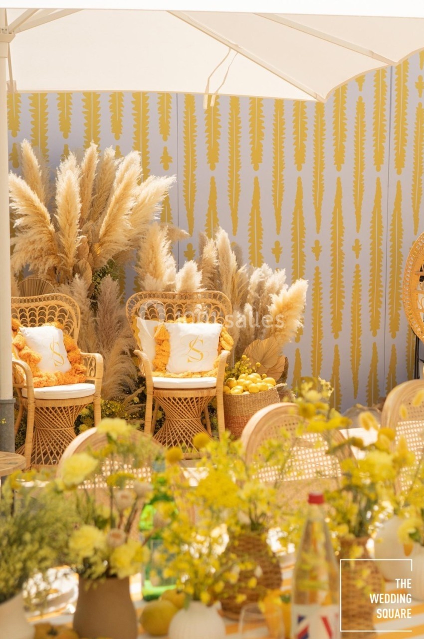 Chaises en rotin entourées de pampas et de décorations jaunes, sous un parasol blanc dans un cadre lumineux.