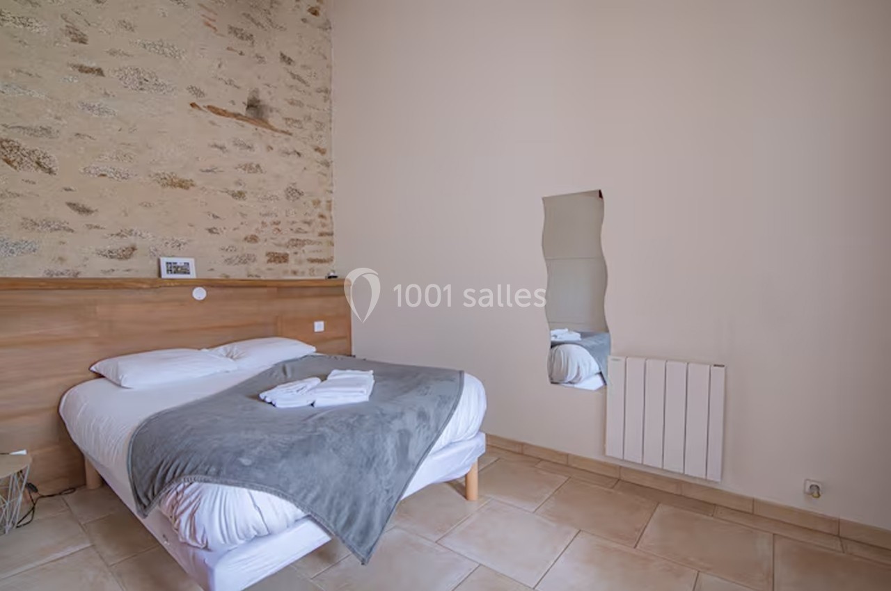 Chambre avec lit double, mur en pierre apparente, miroir mural ondulé et sol en carrelage beige.