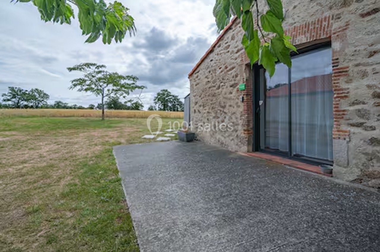 Terrasse en béton attenante à une maison en pierre, donnant sur un jardin avec arbres et champ en arrière-plan.
