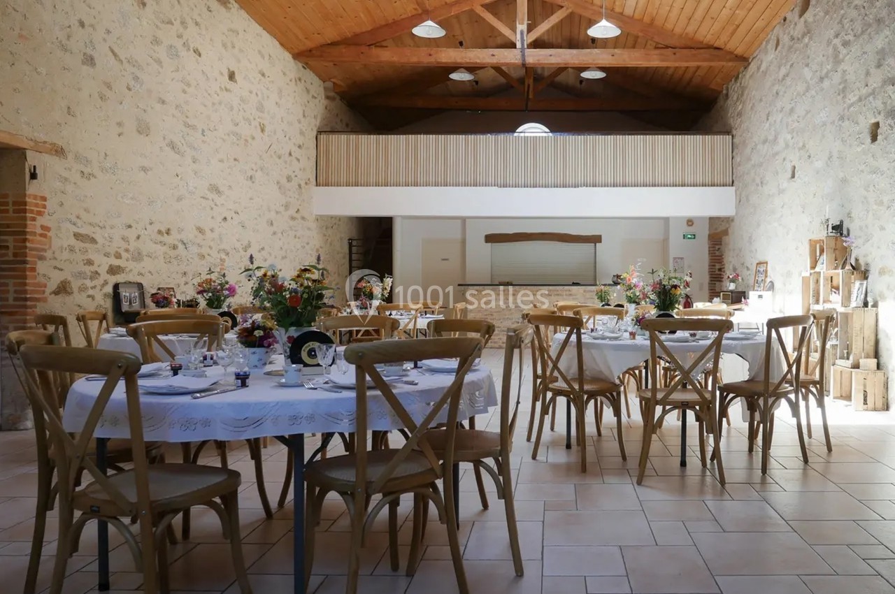 Salle de réception avec murs en pierre, plafond en bois et tables décorées de fleurs pour un événement.
