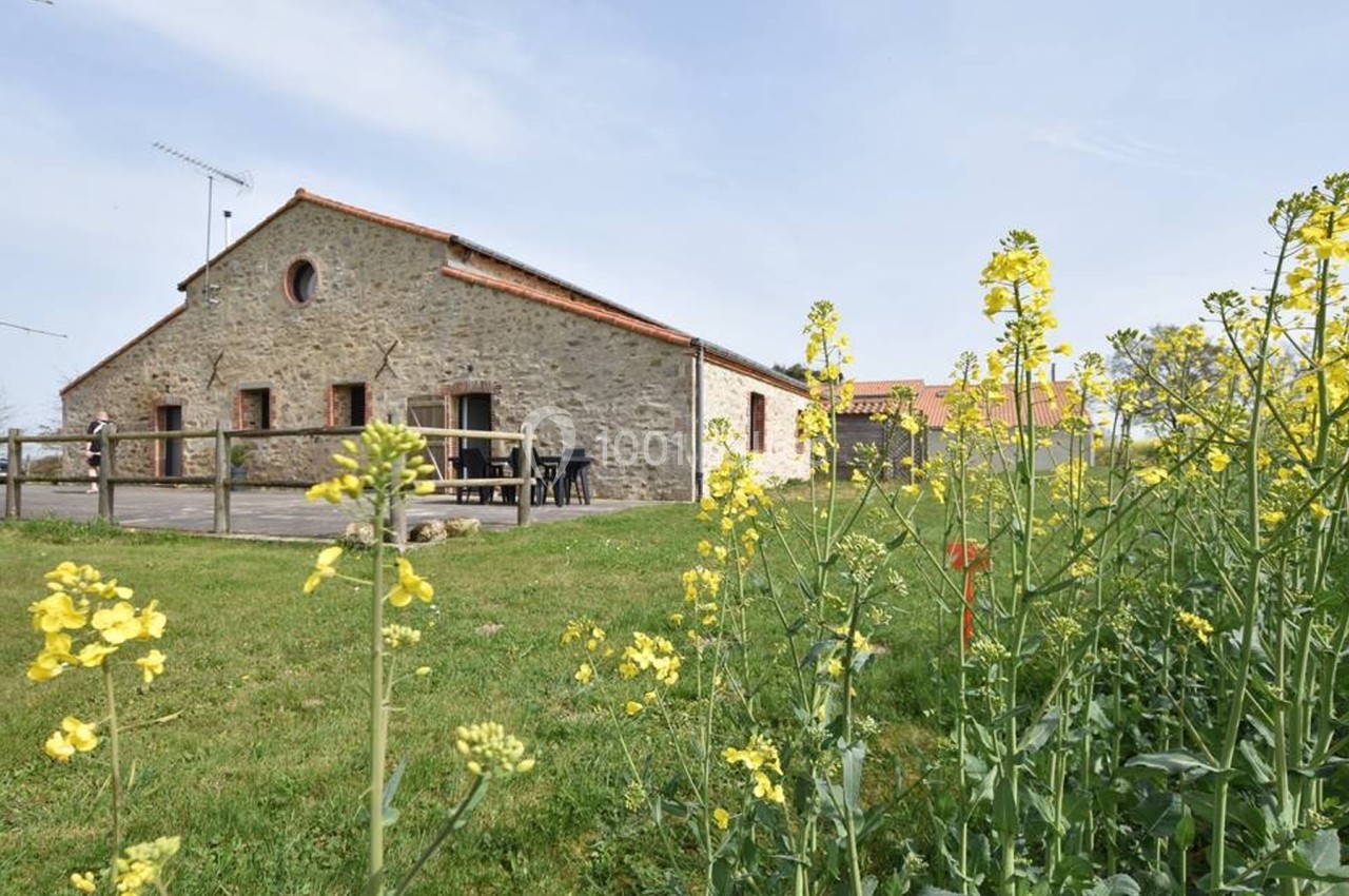Bâtisse en pierre avec toit en tuiles, entourée de verdure et de fleurs jaunes sous un ciel dégagé.