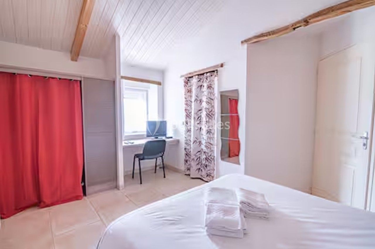 Chambre lumineuse avec lit, bureau près d'une fenêtre, rideaux rouges et imprimés, plafond en bois clair.