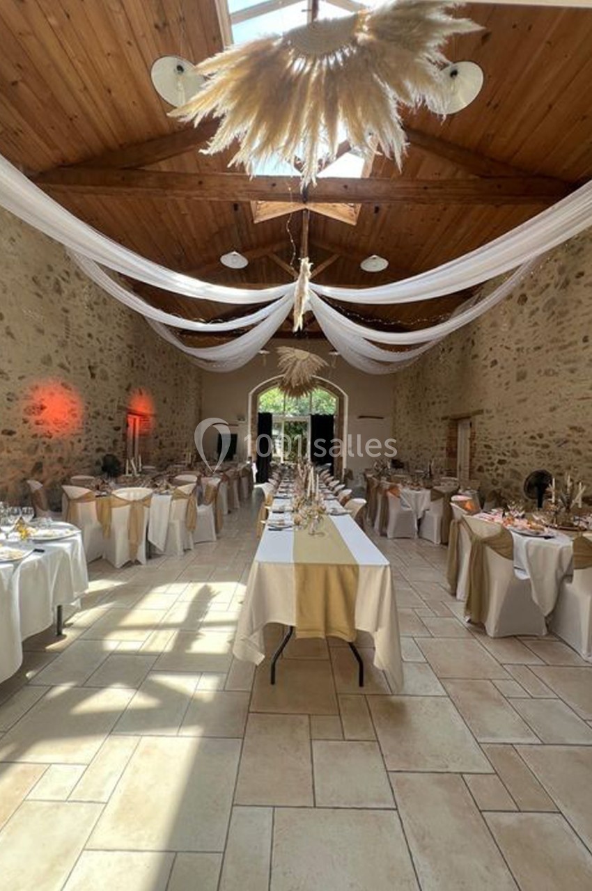 Salle de réception en pierre avec tables dressées, nappes blanches et décorations suspendues sous un plafond en bois.