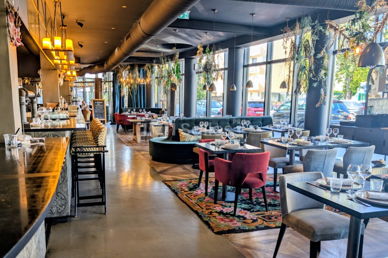 Salle de restaurant aux tables dressées, banquettes et fauteuils rouges et gris, tapis fleuri au sol, bar en pierre sombre à gauche et grandes baies vitrées.