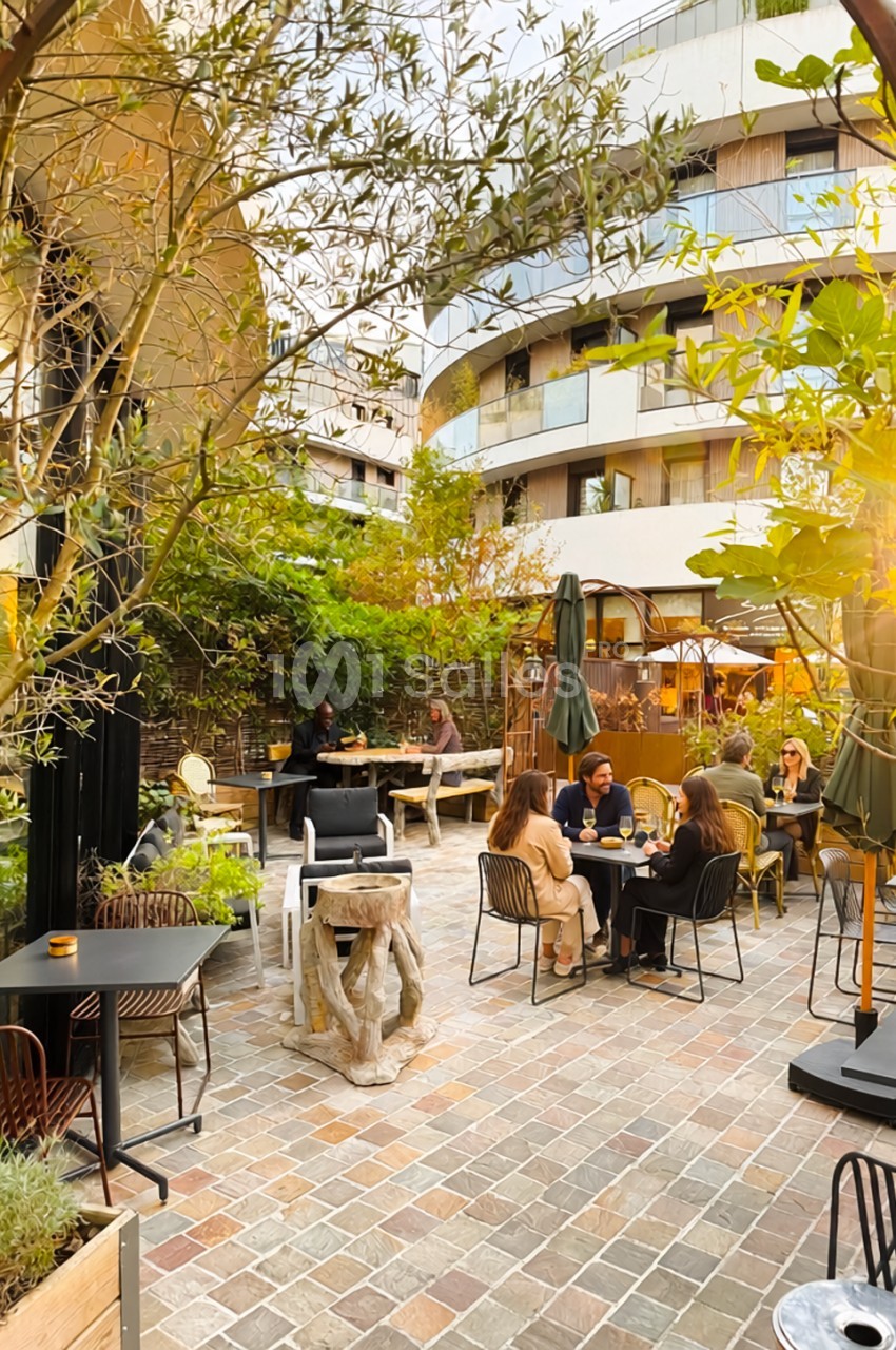 Terrasse pavée et arborée avec tables et chaises en métal, où des convives sont attablés sous des oliviers, devant un immeuble arrondi à balcons.