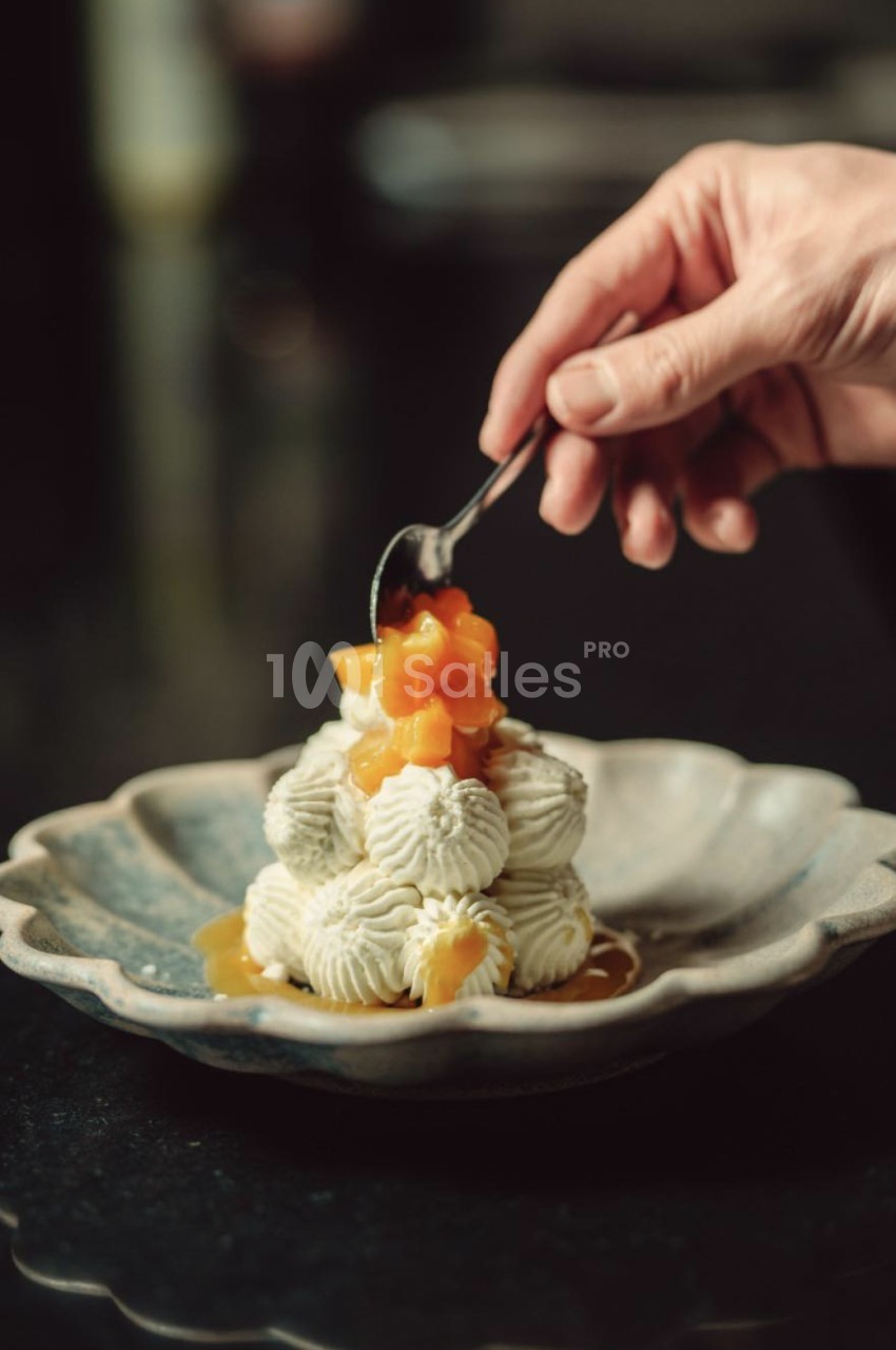 Une main tient une cuillère qui dépose des morceaux de fruit orangé et une sauce dorée sur une pyramide de meringues ou chantilly en rosaces, posée sur une assiette bleutée à bord festonné.