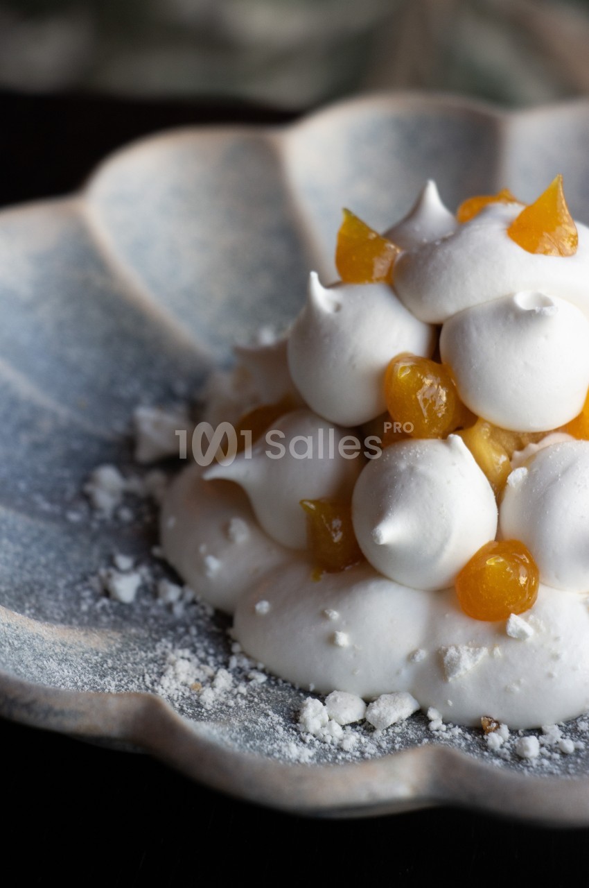 Dessert en dôme de meringue blanche garni de morceaux de fruit confit orangés, posé sur une assiette à bords ondulés et saupoudré de sucre glace.
