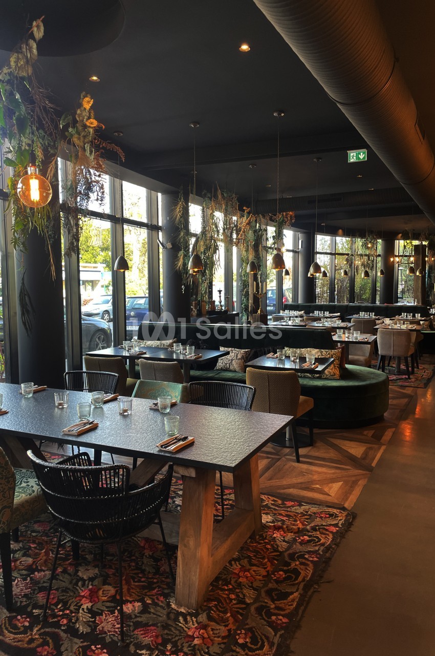 Salle de restaurant aux tables en bois sombre, chaises noires en rotin et banquettes velours vert, avec plantes suspendues et lumière tamisée.